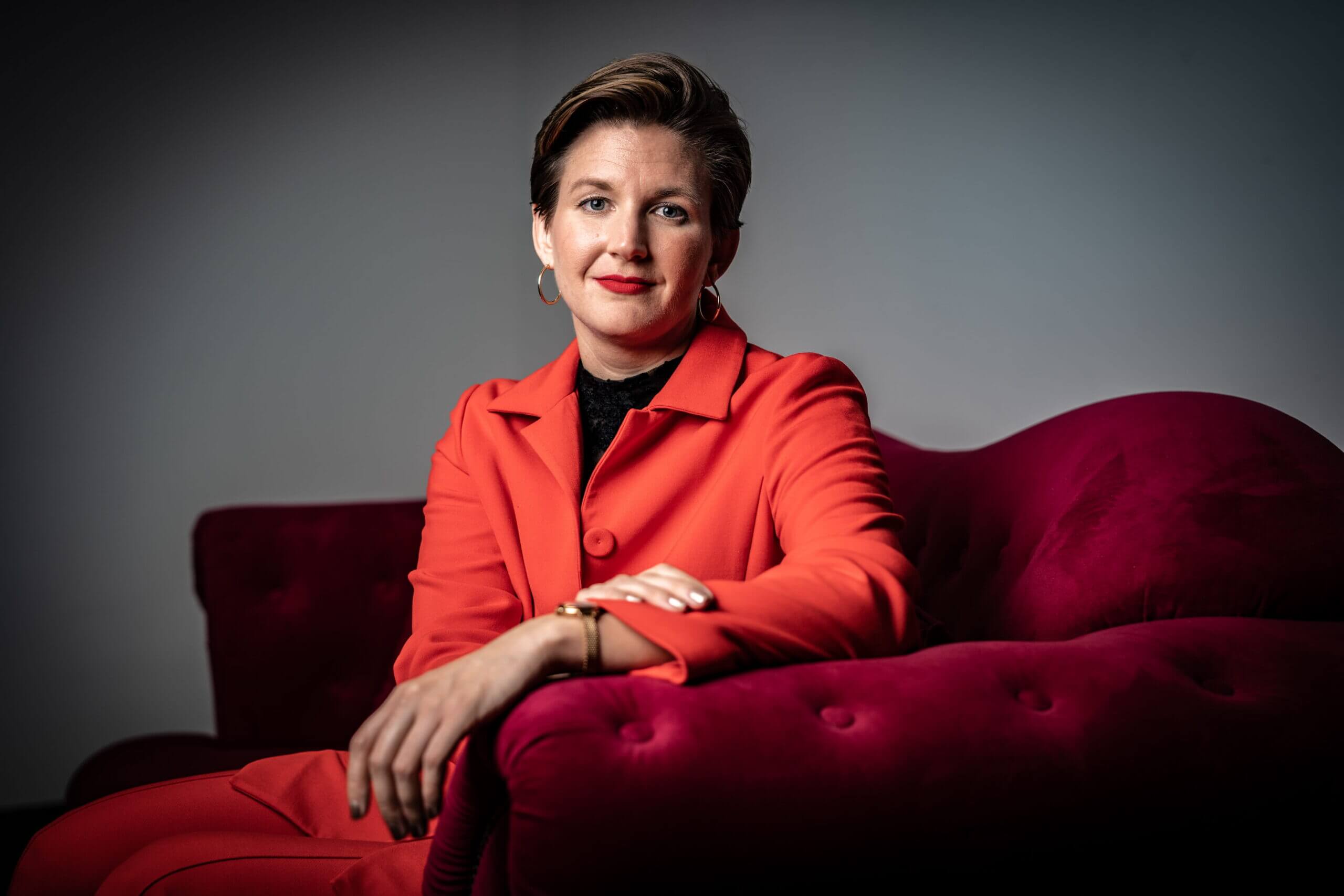 Geschäftsführerin Dr. Simone Burel sitzt auf einem roten Sofa, der Kamera zugewandt. Simone trägt einen roten Hosenanzug, roten Lippenstift und kurzgeschnittene brünette Haare. Sie stützt sich mit einem Arm auf dem Sofa ab und lächelt mit geschlossenem Mund in die Kamera.