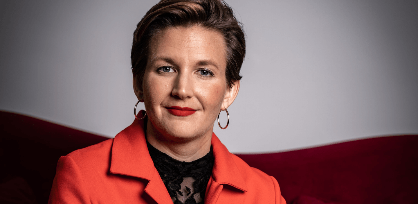 Geschäftsführerin Dr. Simone Burel sitzt auf einem roten Sofa, der Kamera zugewandt. Simone trägt einen roten Hosenanzug, roten Lippenstift und kurzgeschnittene brünette Haare. Sie lächelt mit geschlossenem Mund in die Kamera.