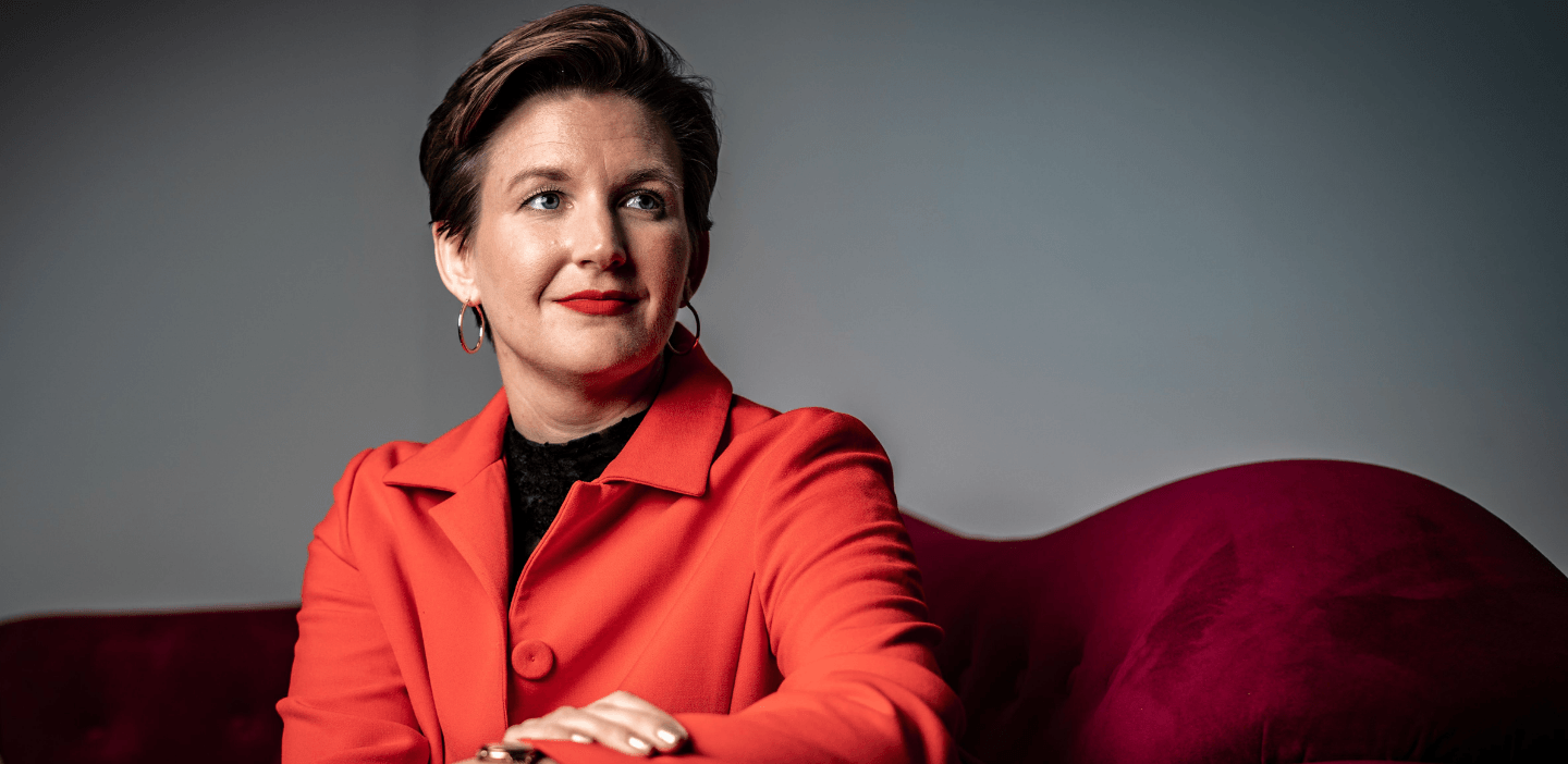 Geschäftsführerin Dr. Simone Burel sitzt auf einem roten Sofa, leicht abgewandt. Sie schaut an der Kamera vorbei schräg nach oben rechts. Simone trägt einen roten Hosenanzug, roten Lippenstift und kurzgeschnittene brünette Haare. Sie stützt sich mit einem Arm auf dem Sofa ab und lächelt mit geschlossenem Mund.