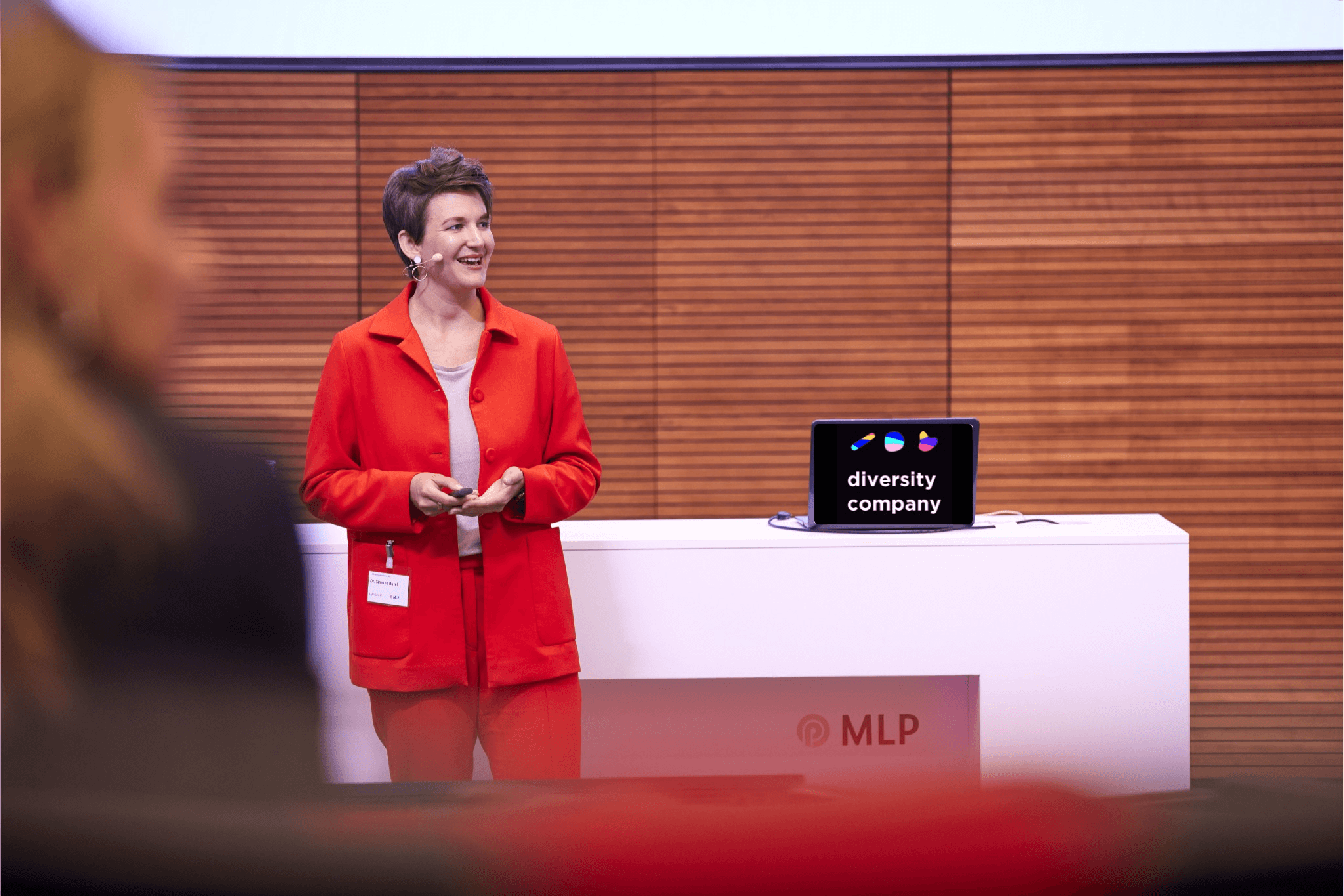 Dr. Simone moderiert eine Veranstaltung beim Finanzdienstleister MLP. Hinter ihr auf einem Tisch steht ein Laptop mit dem diversity company-Logo. Simone trägt einen roten Hosenanzug und kurzgeschnittene brünette Haare.